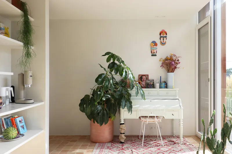 White writing desk with wire chair beside a large potted plant and patterned rug on terracotta tiles