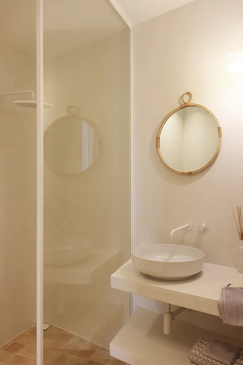 Bathroom vanity with vessel sink and round rattan mirror beside a glazed shower enclosure