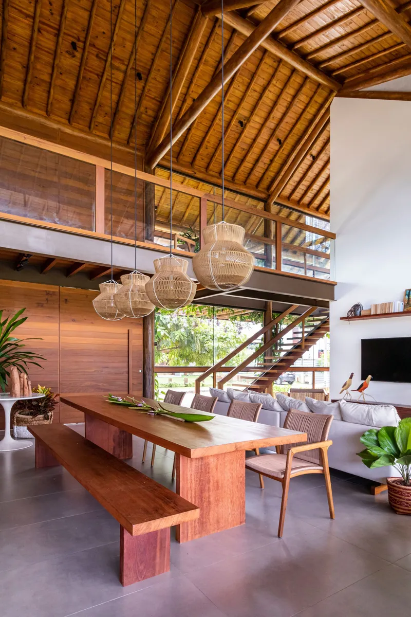 Timber dining table and bench beneath woven pendant lights with mezzanine and pitched ceiling above