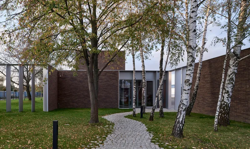 Cobblestone pathway leading through birch trees toward a courtyard entrance between brick volumes