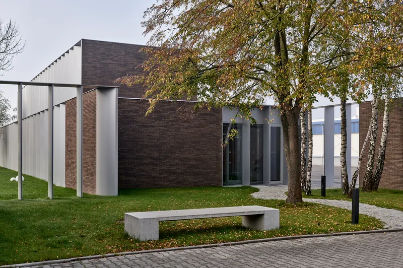 Dark brick facade with vertical metal and glass openings framed by birch trees on a grass lawn