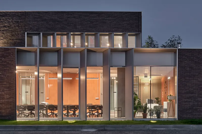 Double-height glazed facade with deep recessed bays flanked by dark brick walls at twilight