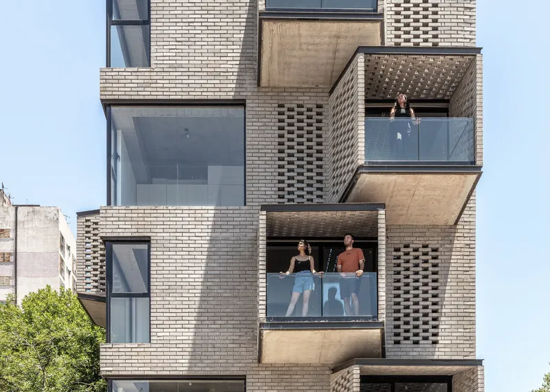 Stacked brick facade with projecting balconies, perforated screens, and residents looking out under clear midday sky