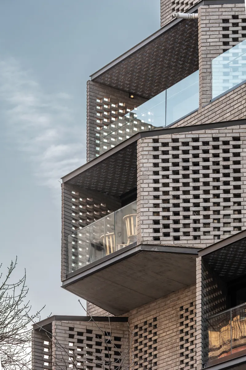 Upward view of cantilevered balconies clad in perforated brick with glass railings under overcast sky