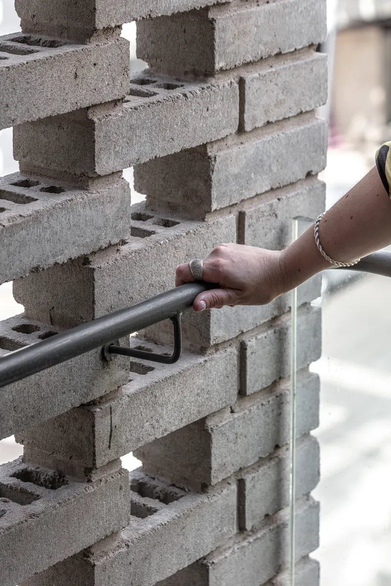 Hand gripping a metal railing mounted to a perforated brick wall in bright daylight