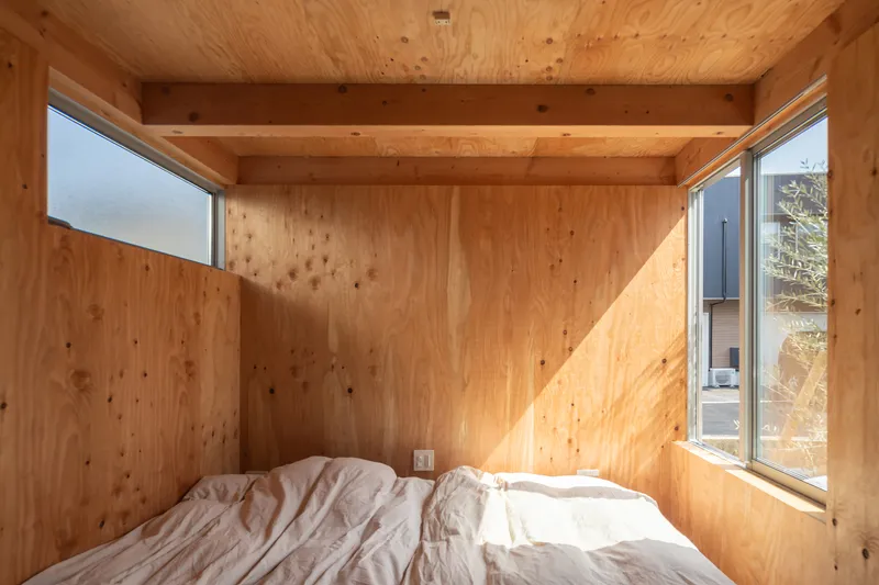 Plywood-lined bedroom with exposed beams and corner windows casting sunlight onto a white futon