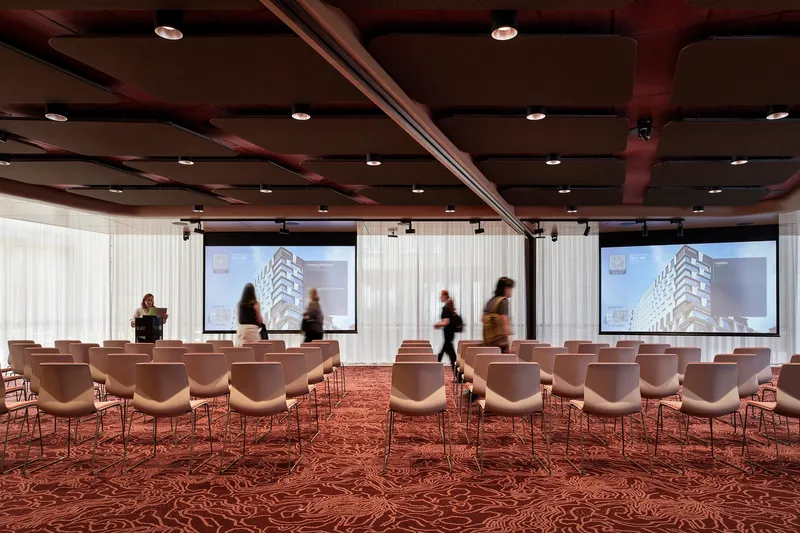 Conference room with patterned burgundy carpet, white chairs and people moving past projection screens beneath dark timber ceiling