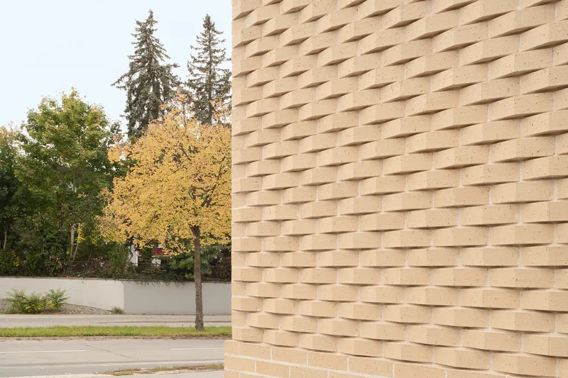 Textured brick facade with projecting headers beside a young tree in autumn leaf