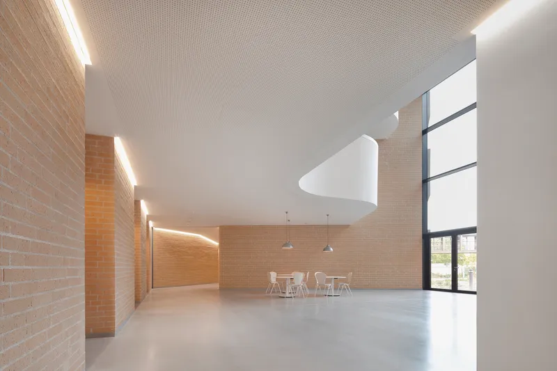 Double-height gathering space with yellow brick walls, perforated ceiling, and tall windows overlooking trees