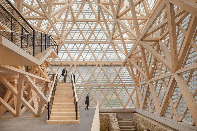 Interior atrium with diagonal timber lattice structure and wooden staircase above excavated stone foundations