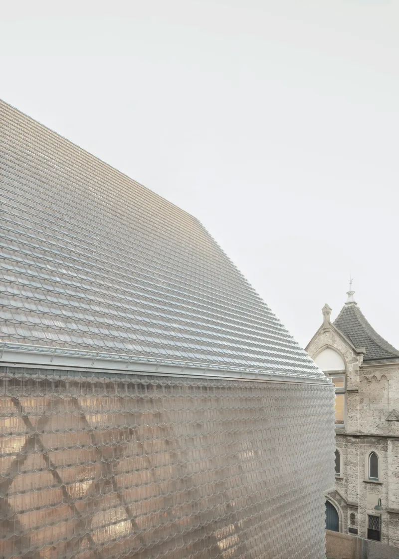 Faceted glass tile roof volume adjacent to a historic stone church tower under grey skies