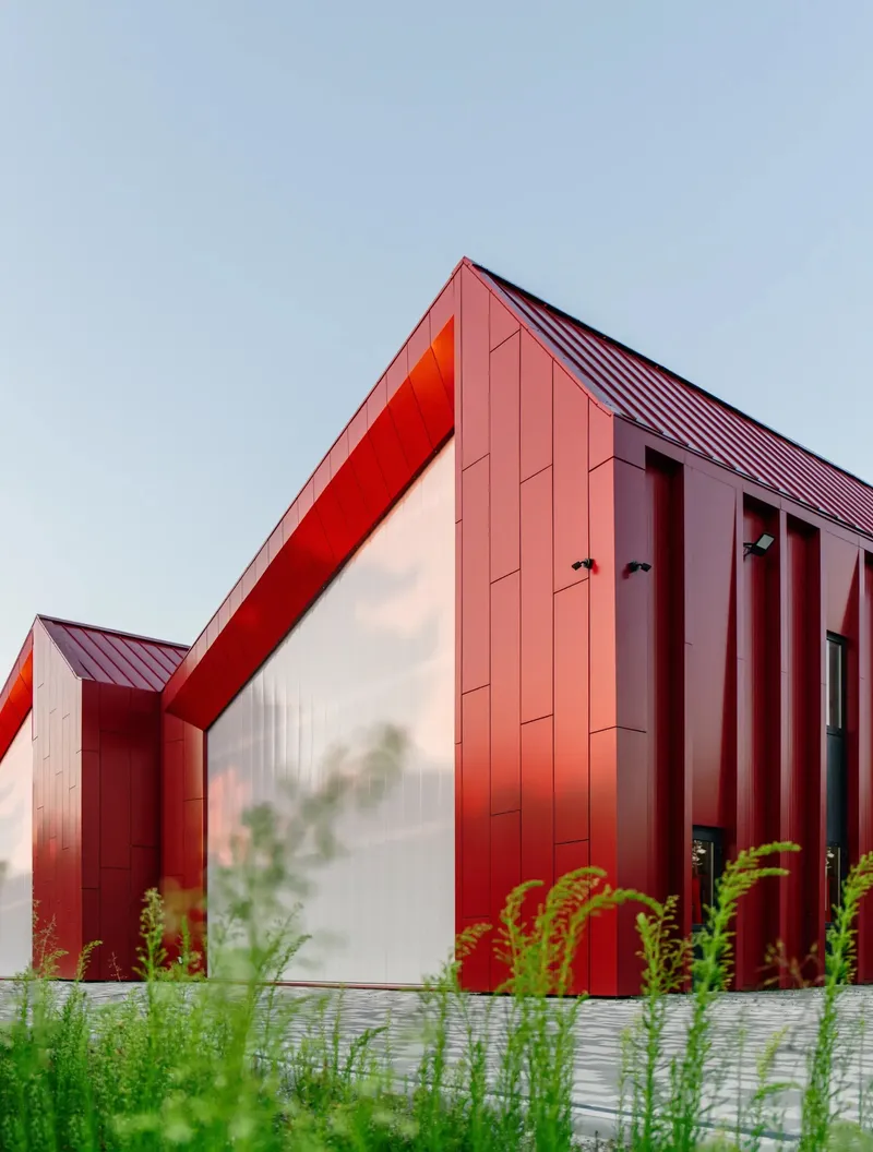 Angled view of red metal facade with recessed white entry framed by vertical fins and grasses