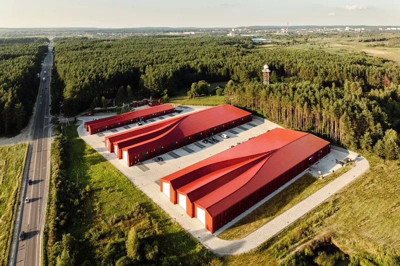 Aerial view of three red metal storage buildings with sawtooth roofs surrounded by forest and farmland