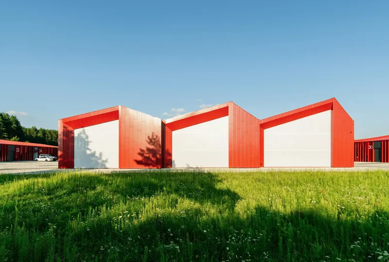 Three linked warehouse volumes with white end walls and red metal roofs beside a grassy field