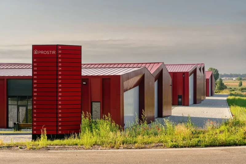 Red locker tower and timber-clad entry volumes facing a gravel roadway at dusk