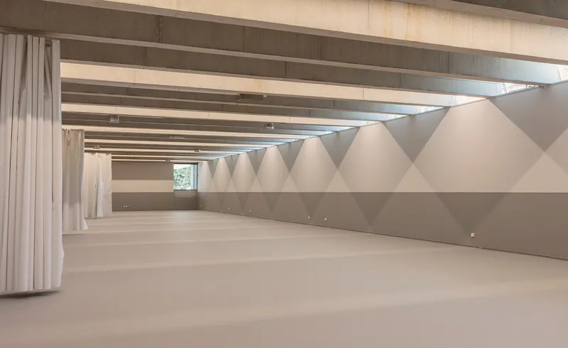 Long corridor with faceted white wall panels and exposed timber beams beneath a concrete ceiling