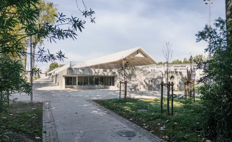 Entrance pathway framed by bamboo and young trees leading to a glass-fronted pavilion with a gabled concrete roof