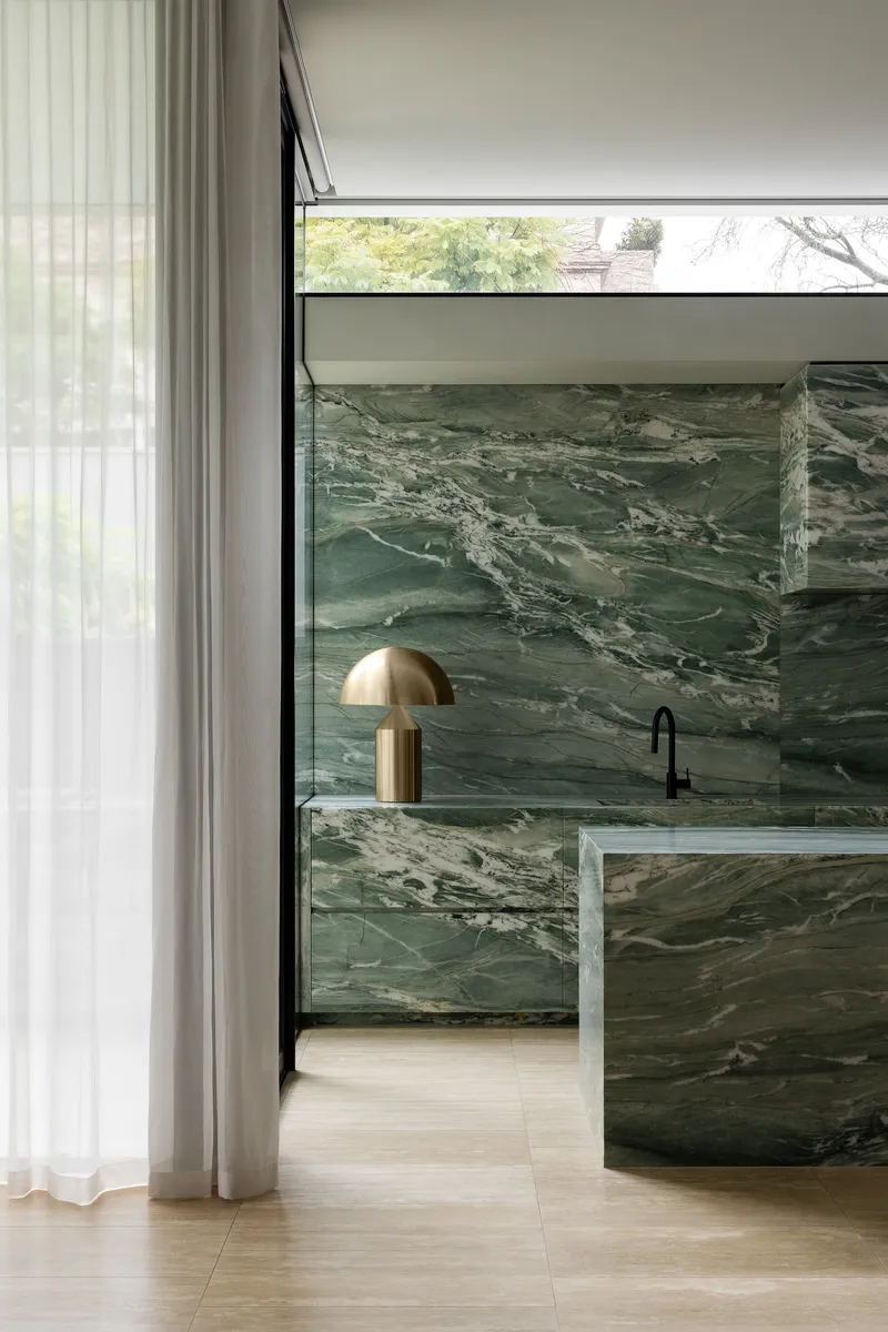 Kitchen island clad in green veined marble with a brass table lamp beside sheer curtains