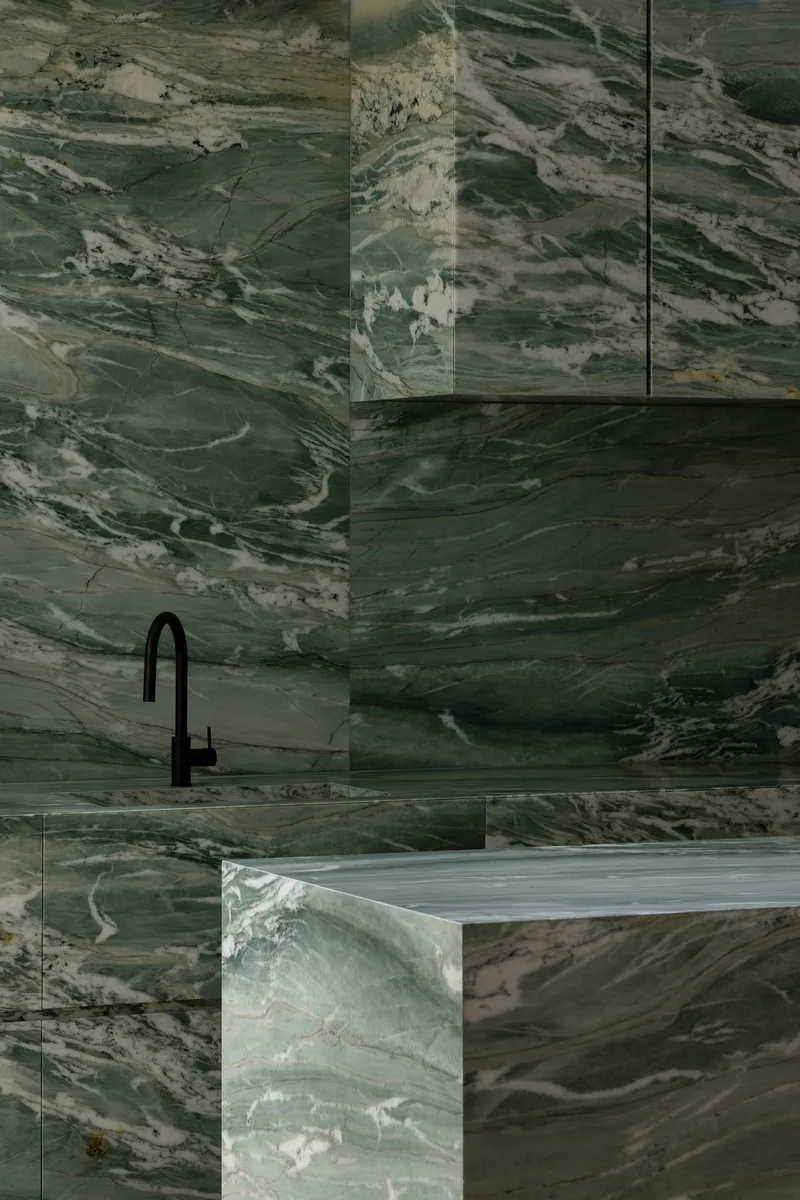 Corner detail of green veined marble surfaces meeting around a black tap fixture and rangehood