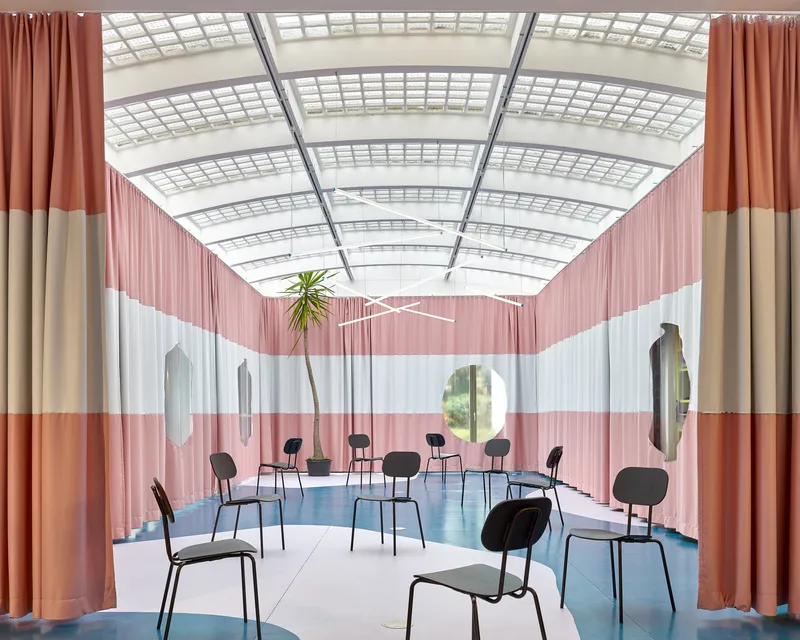 Interior hall with black chairs on curved blue flooring beneath a gridded skylight and striped curtains