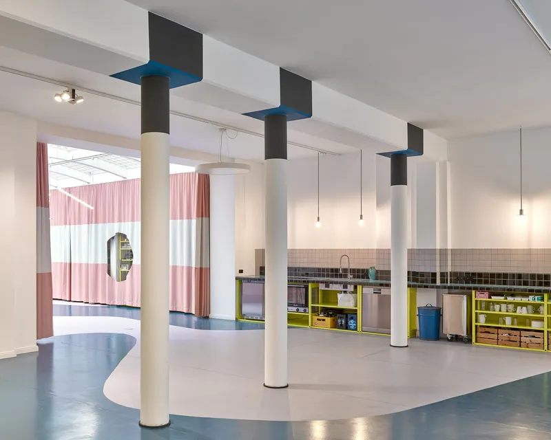 Open space with cylindrical columns capped by blue squares looking toward a yellow kitchen unit