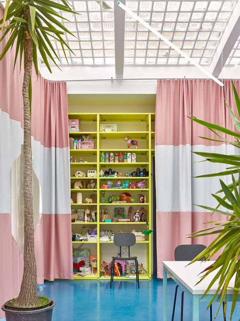 Yellow shelving unit filled with toys behind parted striped curtains and potted palm fronds