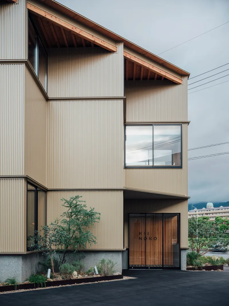 Front elevation showing corrugated metal walls and timber entrance doors with planted foreground under cloudy skies