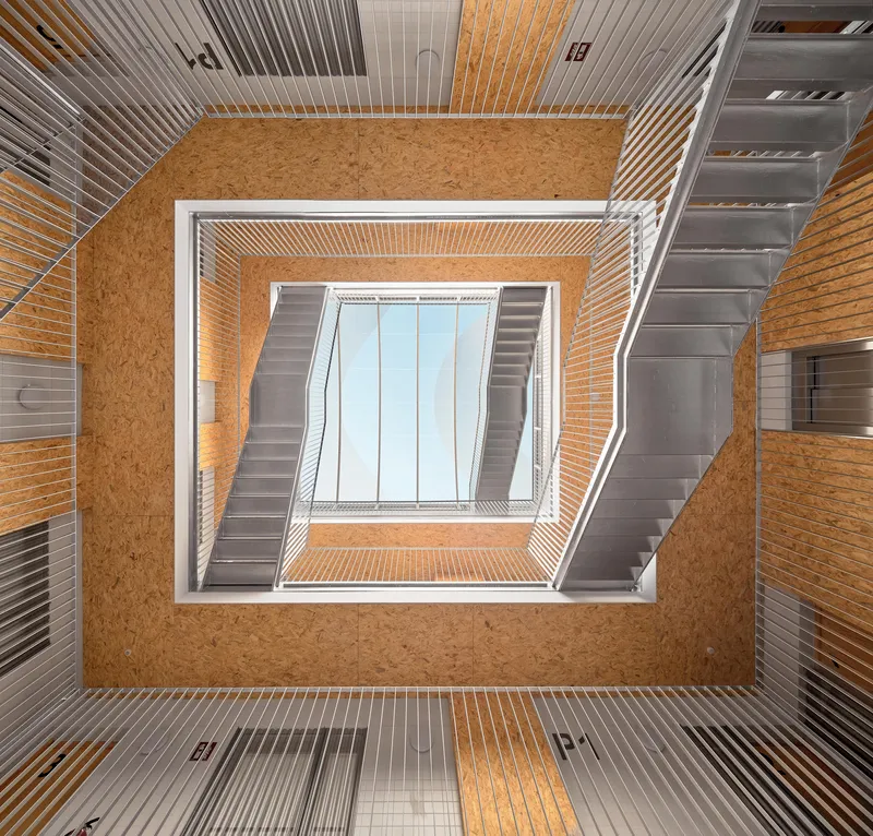 Looking up through a square stairwell with oriented strand board walls and ribbed metal ceilings