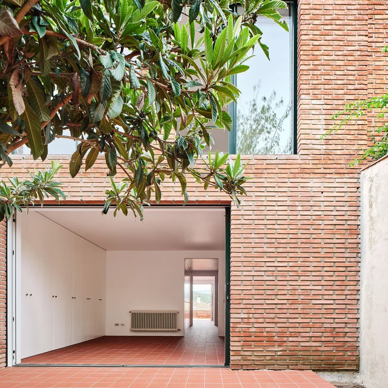 Brick facade with large opening revealing interior corridor and terracotta floor beneath overhanging tree branches