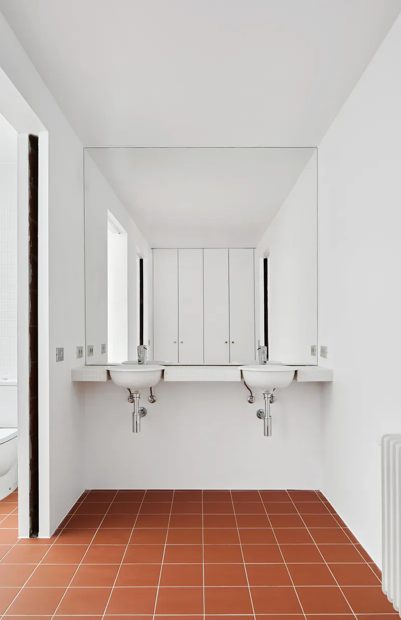 White bathroom with terracotta tile floor, wall-mounted twin sinks and large mirror reflecting cabinet doors