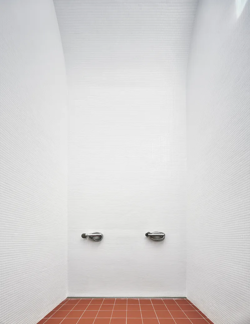 Narrow shower alcove with white mosaic tile walls and two chrome faucets above terracotta floor