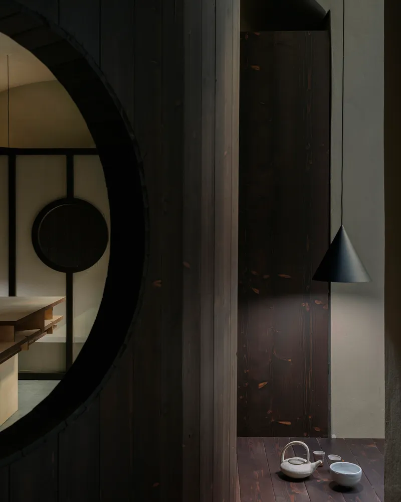 Dark timber wall with circular portal opening and conical pendant light above ceramic tea set