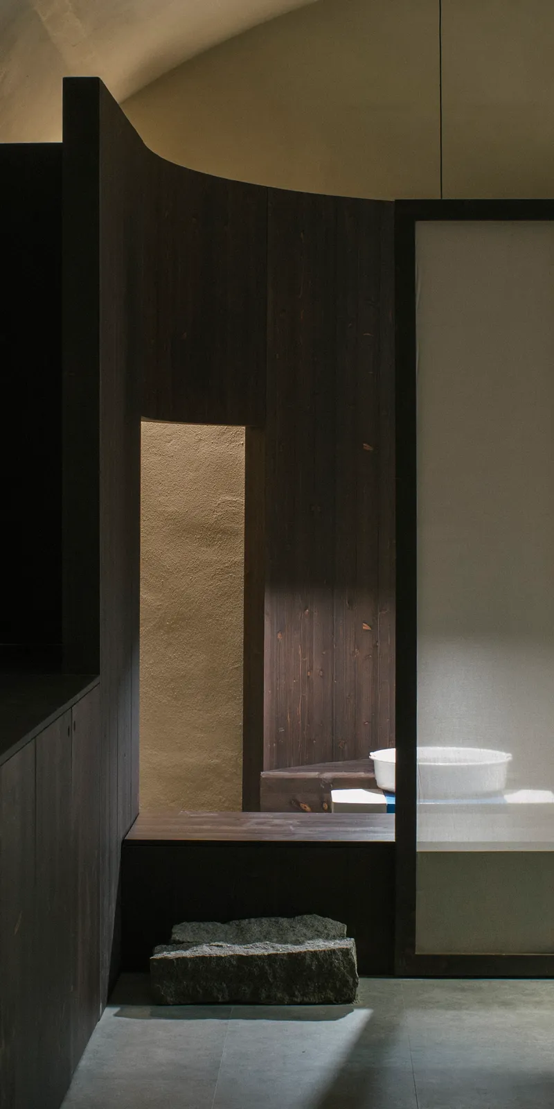 Layered timber partitions with narrow aperture revealing stone basin and shelving beyond