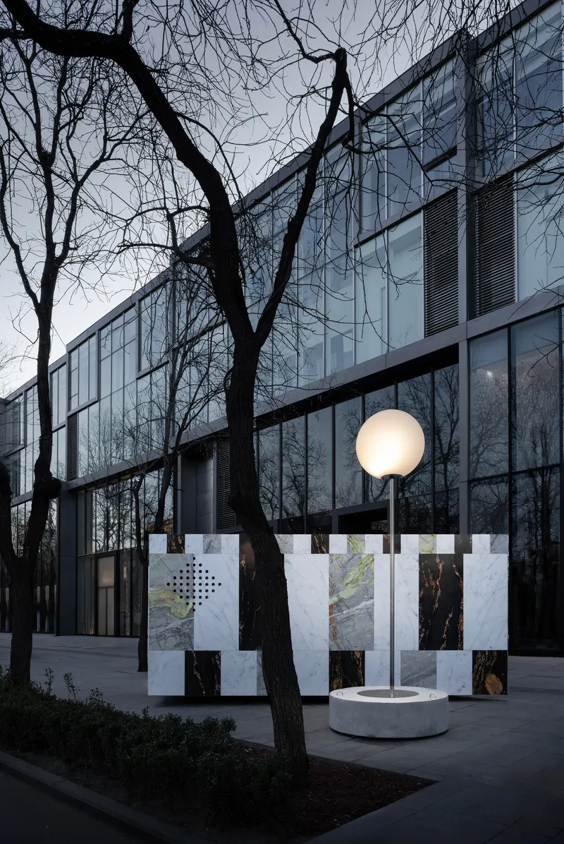 Glass facade behind bare winter trees with a stone-clad installation and illuminated globe lamp at dusk