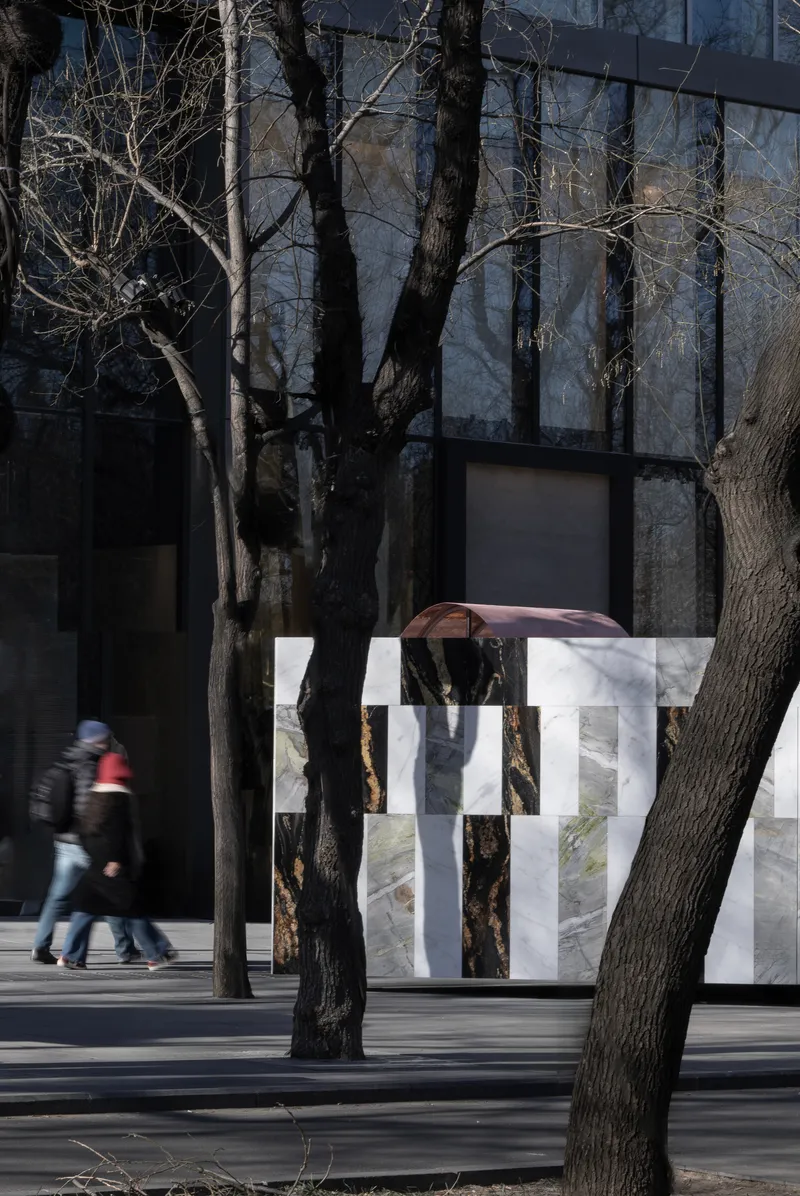 Striped stone wall installation with copper arch element as pedestrians pass beneath bare tree shadows