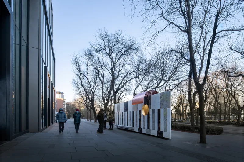 Pedestrians walking along paved plaza with freestanding stone wall and copper tube sculpture at sunset
