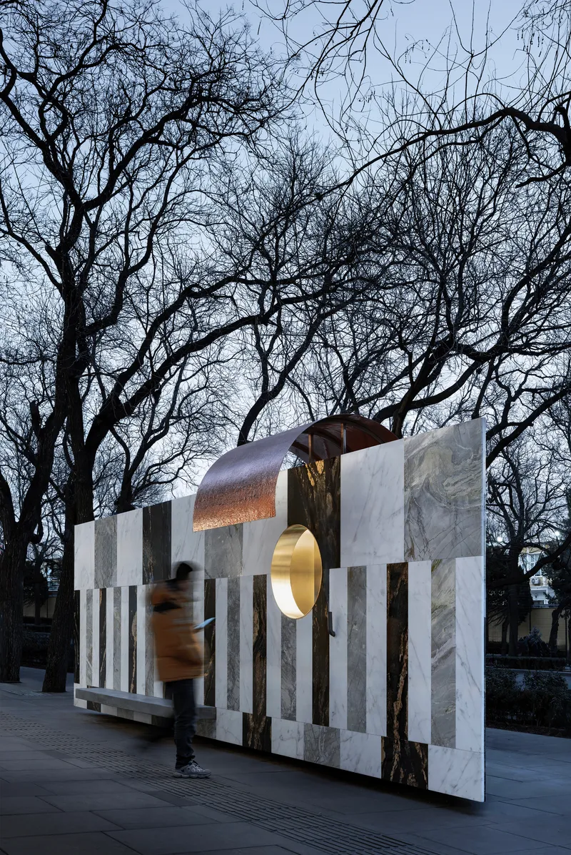 Freestanding wall of vertical stone stripes with copper canopy and pedestrian passing through at twilight