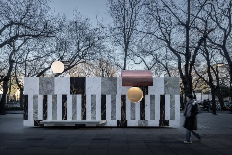 Freestanding pavilion with alternating marble panels and copper canopy in a public plaza with bare winter trees