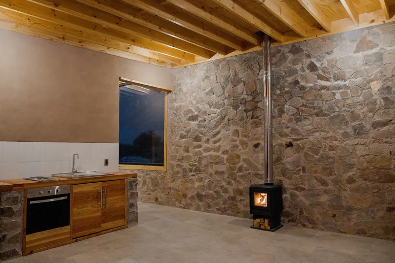 Interior kitchen space with stone wall, timber cabinetry, and freestanding wood stove at dusk