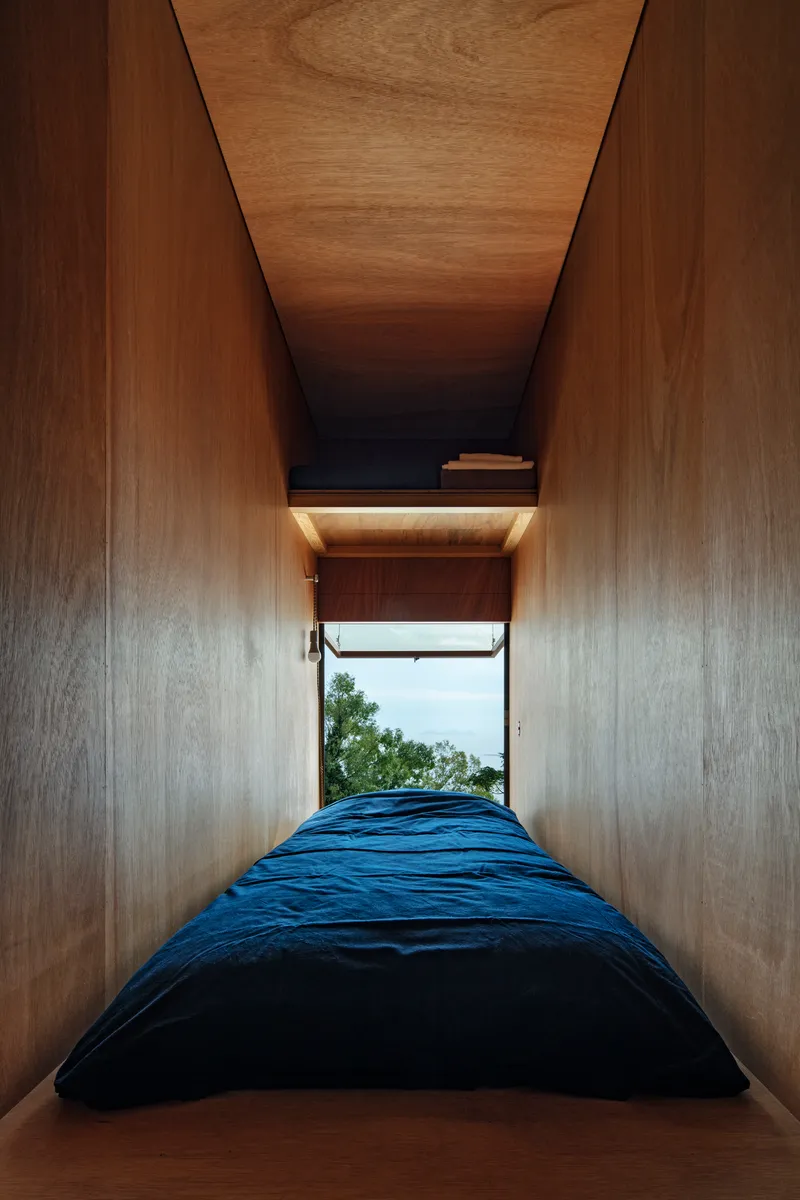 Narrow sleeping nook with plywood walls and ceiling opening to a window with ocean view