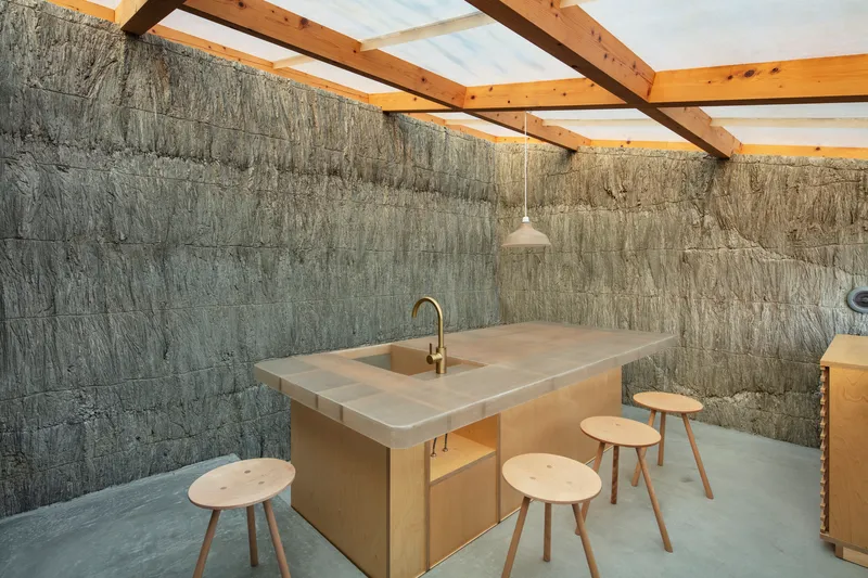 Kitchen island with wooden stools beneath exposed timber roof structure and textured concrete walls