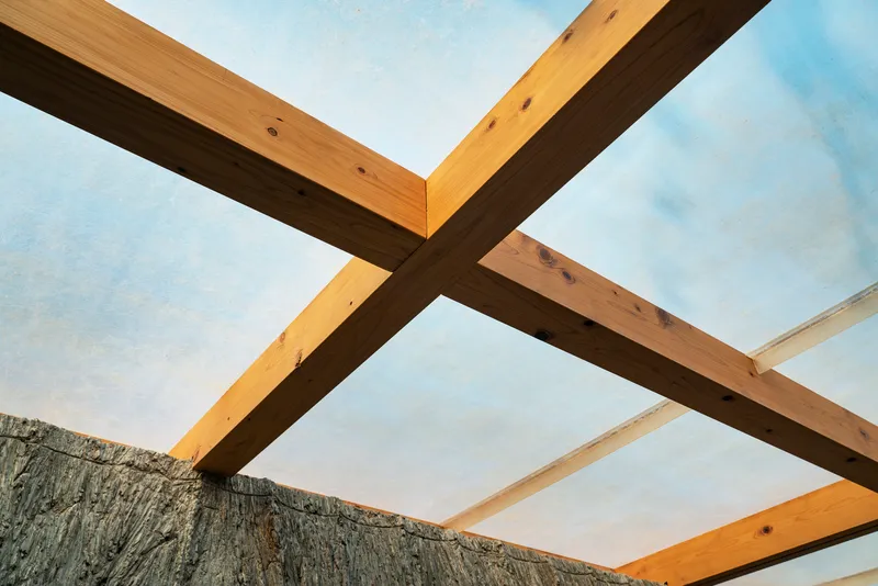 View upward through crossing timber beams framing the hempcrete ceiling against blue sky