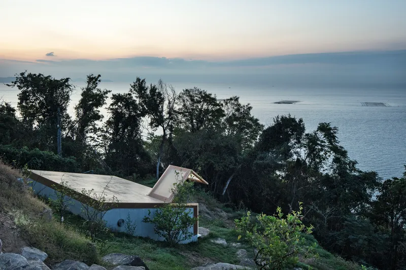 Folded concrete roof nestled into the hillside vegetation above the ocean at dusk