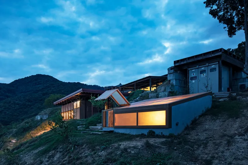 Cluster of volumes with angled roofs and illuminated glazing on the sloped site at twilight