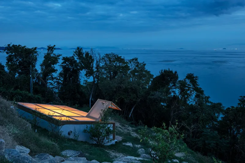 Cantilevered roof illuminated from within overlooking the forested coastal hillside at night