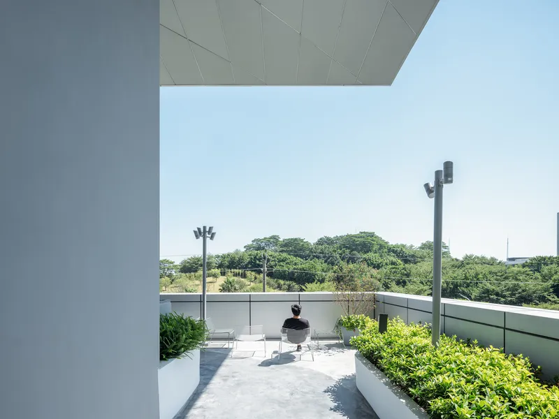 Rooftop terrace framed by projecting slabs with seated figure among planted beds under clear sky