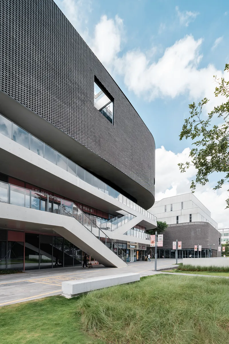 Cantilevered concrete bands extending over glazed entry beneath a dark perforated metal screen with blue sky