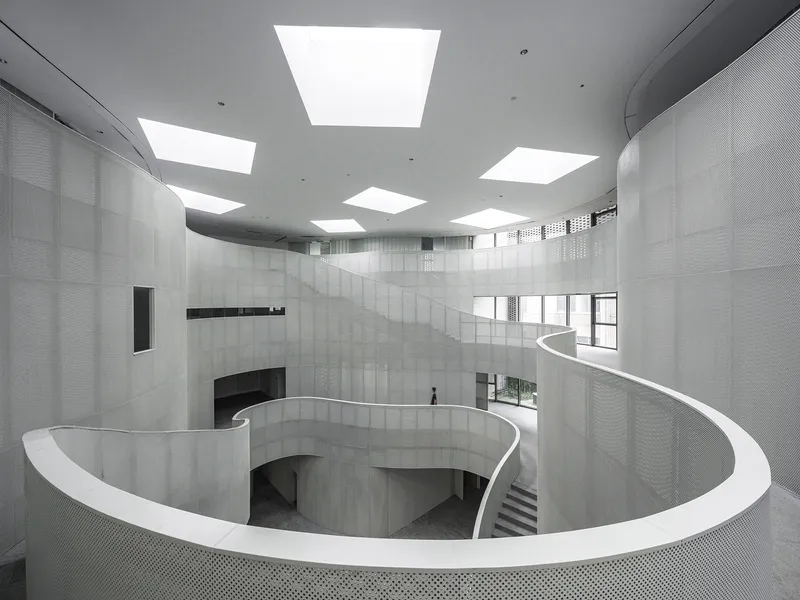 Spiraling white balustrade and concrete walls beneath square skylights in a multi-story atrium with a lone figure