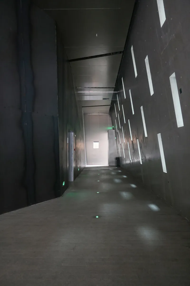 Dark corridor with vertical light slots piercing the concrete wall casting geometric shadows on the floor