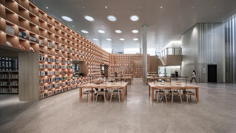 Library interior with timber bookshelf wall, reading tables and a person walking past translucent curtains